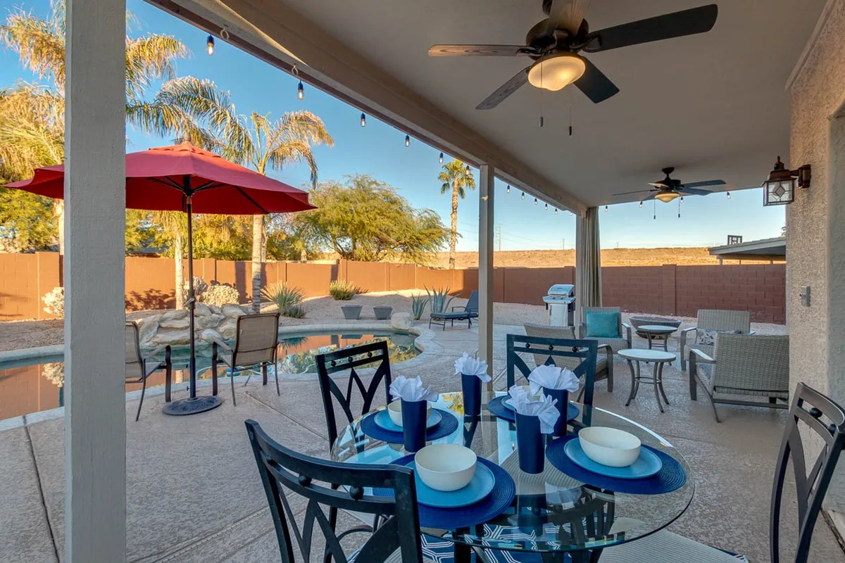 An outdoor dining table is elegantly set beside a glistening pool, shaded by a red umbrella and flanked by swaying palm trees. The area is inviting and cheerful, ideal for alfresco meals or social gatherings by the water.