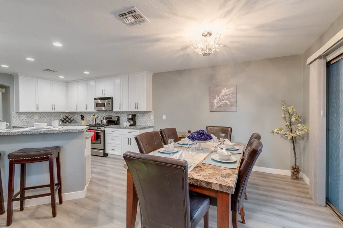 A cozy interior view blending the dining area and kitchen. The dining table is set for a meal, with a chandelier overhead and the kitchen visible in the background, highlighting a harmonious space for sharing meals, conversations, and celebrations.