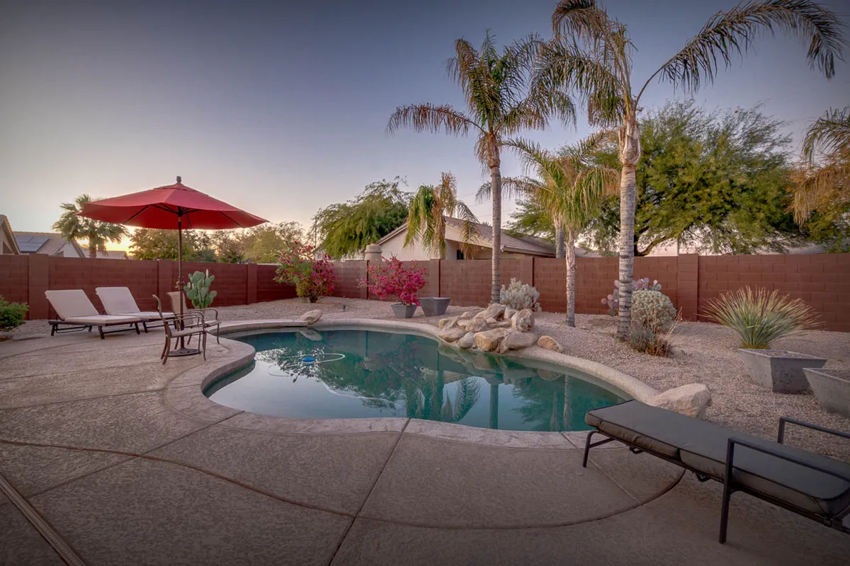 A sunlit backyard with a swimming pool bordered by palm trees, lounge chairs, an umbrella, and lush flowering plants. The space exudes a calm, inviting vibe, perfect for socializing or quiet moments surrounded by nature.