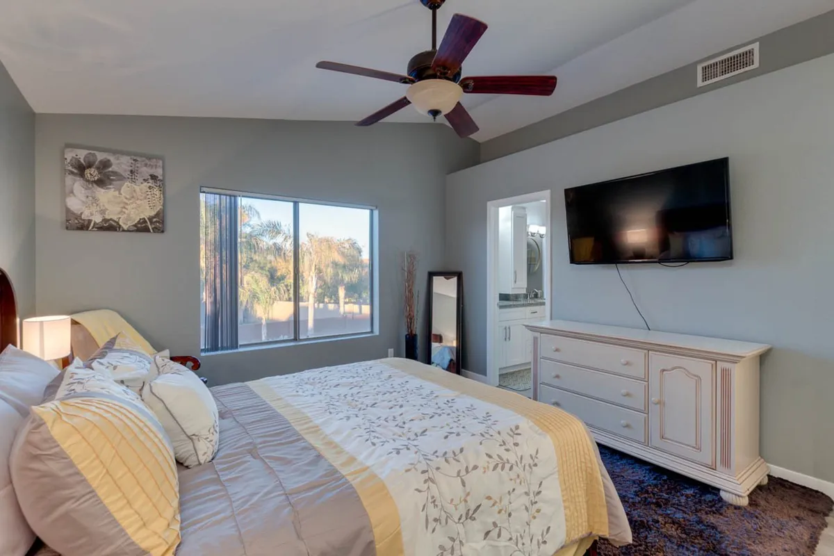 This image captures a serene bedroom scene featuring a bed with decorative pillows, a large window letting in abundant natural light, a TV mounted on the wall, a decorative mirror, and a dresser. The mood is calm and comfortable, ideal for relaxation or unwinding with a movie.