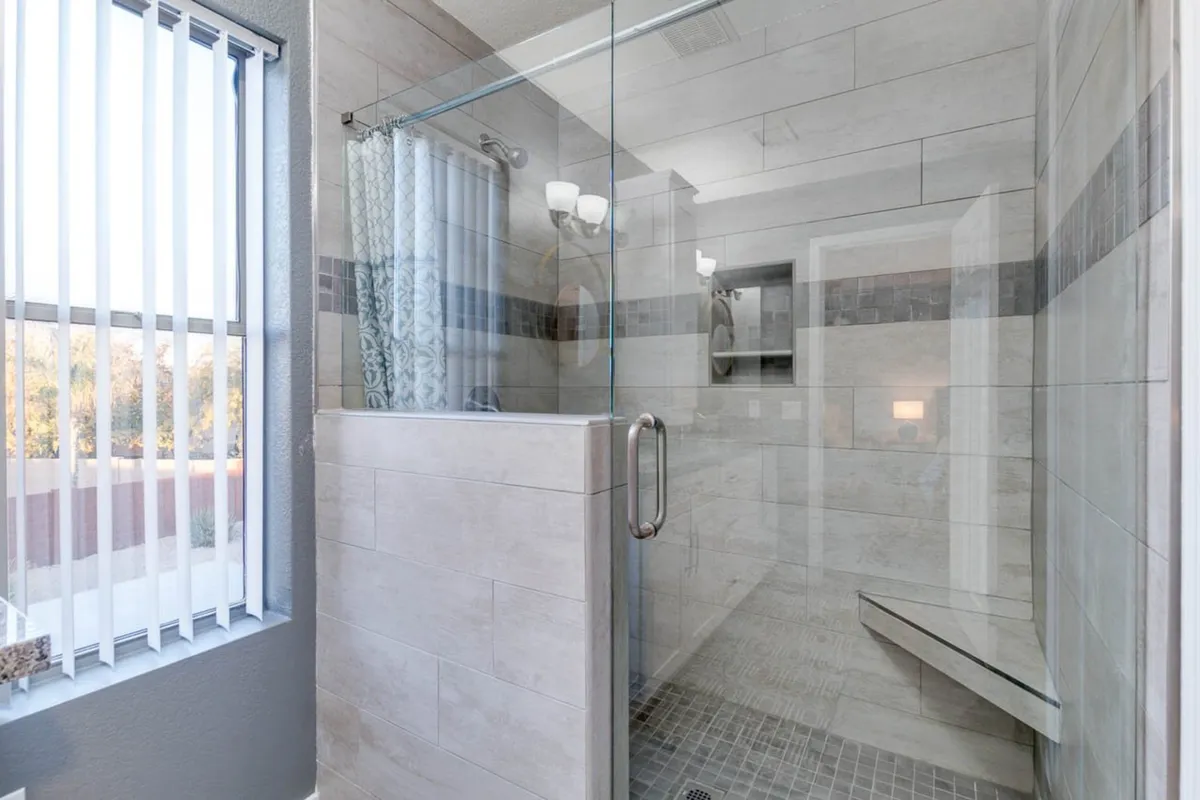 A luxurious, modern glass shower enclosure with textured tile walls, stylish fixtures, and soft daylight filtering through vertical blinds. The spa-like ambiance invites relaxation and unwinding after a day of exploration.