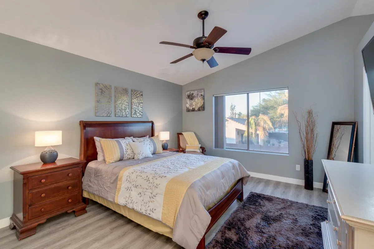 A serene and spacious bedroom featuring a comfortable bed flanked by nightstands, a large window letting in natural light, and a ceiling fan overhead. The room's soft lighting and calming décor make it ideal for quiet relaxation.