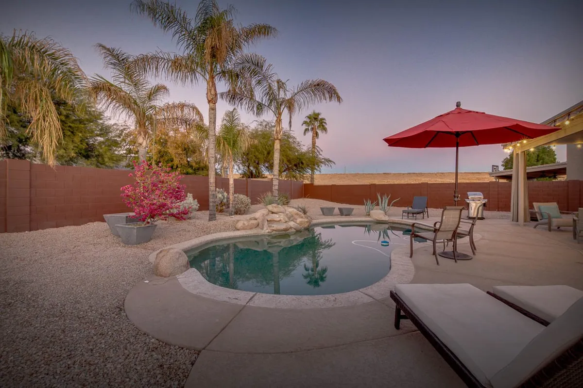 A beautifully landscaped backyard features a sparkling swimming pool surrounded by palm trees and rock features. Several lounge chairs are arranged beside the water, with a vibrant red umbrella adding a pop of color. The mood is serene, perfect for unwinding by the pool or enjoying a quiet moment with nature.