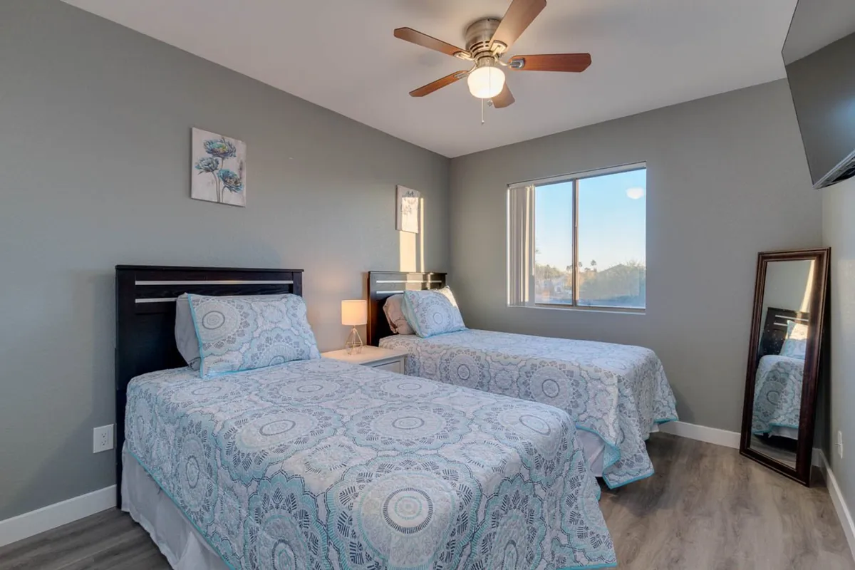 This image displays a guest bedroom with two twin beds with patterned bedding, a large window providing natural light, decorative wall art, and a mirror reflecting the room. The atmosphere is comfortable and cozy, perfect for families or friends sharing the space.